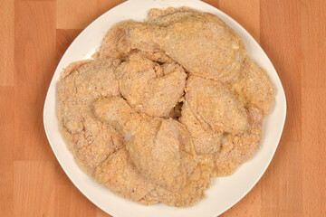 Several pieces of breaded raw chicken, including drumsticks and cutlets, are arranged on a white plate ready for cooking
