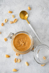 Glass jar  with creamy peanut butter with spoon on grey background with scattered roasted nuts