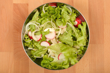 An overhead shot shows a stainless steel bowl filled with a vibrant spring salad of crisp green lettuce, thinly sliced white onions, and red and white radish slices on a wooden surface