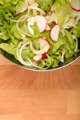 An overhead shot shows a stainless steel bowl filled with a vibrant spring salad of crisp green lettuce, thinly sliced white onions, and red and white radish slices on a wooden surface