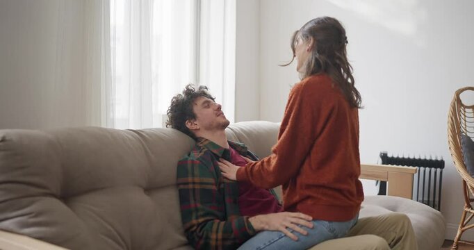 Happy brunette girl sits on her boyfriend's lap who is relaxing on the couch during a fun romantic scene in a bright apartment at home