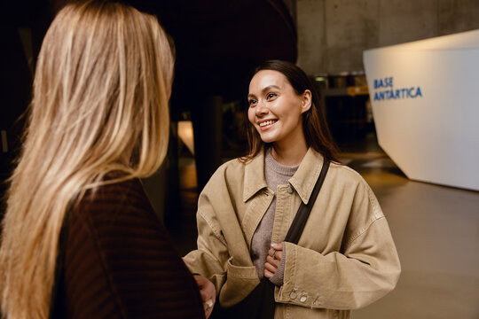 Two young women engage in friendly conversation indoors near a modern exhibit marked "Base Antarctica," smiling warmly. A concept of connection, shared curiosity, and the spirit of exploration.