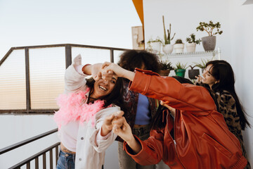 A group of joyful young women of different ethnicities in festive clothes dance and laugh together on a balcony at sunset, surrounded by potted plants. Concept of girls' party and friendship.