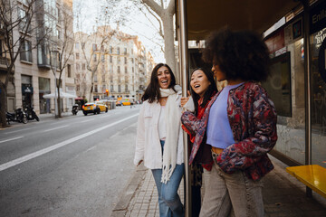 Three stylish friends share a laugh at a city bus stop, enjoying each other's company on a quiet urban street with yellow taxis and bare trees. Concept of waiting for a bus with girlfriends.
