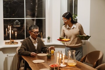 At a warm evening dinner, an Asian woman serves food to an elderly Asian man at a candlelit table. Concept of a peaceful and intimate family moment, a couple's dinner.