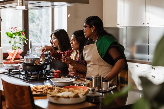 A group of young Indian women is cooking in a bright kitchen, preparing dinner, wearing aprons and traditional attire, smiling, and enjoying the cooking process with fresh ingredients and utensils.