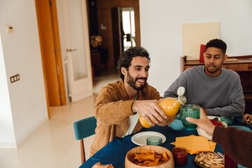 A cheerful man pours orange juice into a friend's glass while sitting at a table full of snacks, and his male guest watches with a smile. Concept of a relaxed atmosphere of the gathering with friends.