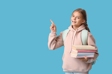 Cute schoolgirl with books and backpack pointing at something on blue background
