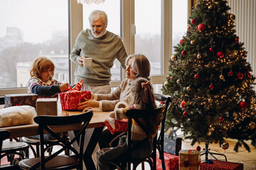 A senior White man, his wife, and their grandson are opening Christmas gifts together by the decorated tree, sitting around a table in a cozy, well-lit room with festive decorations.