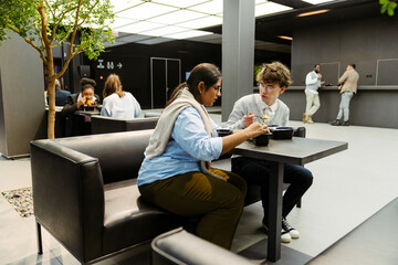 An Indian woman and her White male colleague eat lunch from black lunch boxes on a black sofa in...