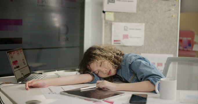 Exhausted young business woman graphic designer sleeping on desk after long hours of work. Overworked, tired and burnout female falls asleep at messy table with laptop after long workday. nap at work.