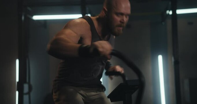 Close up of a male athlete with a shaved head in a tank top doing an intense workout and fast movement on an exercise bike in a modern gym