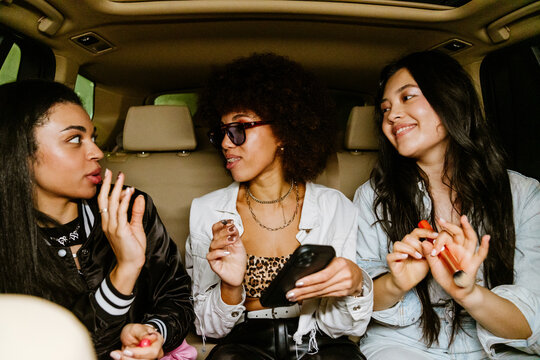 Three mixed-race women laughing and chatting in the backseat of a car while doing their makeup and holding drinks, capturing a spontaneous and joyful moment on the way to a night out.