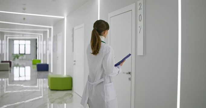 Rear view of female doctor in white coat holding tablet in hands walking down corridor and entering needed office during work in hospital