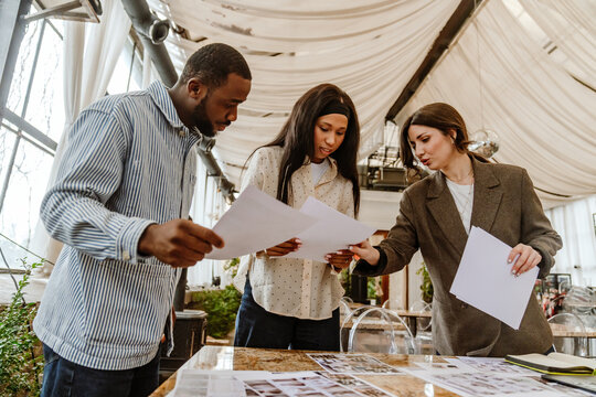 A young Black groom and bride are discussing printed materials with a young White female wedding planner in a decorated event venue filled with natural light and draped fabric ceiling.