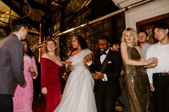 A multicultural group of adult wedding guests dances indoors with the bride in a white gown and the groom in a black suit during a joyful evening wedding celebration filled with formal outfits.