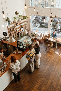 Adults inside a modern cafe with wooden floors and large windows, some standing in line to order from the barista at the counter while one man sits by the window enjoying a drink.