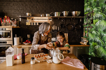 A school-aged White girl wearing a multicolored sweater, helping her elderly mid-70s grandma mix some dough in a metal bowl as they stand in a modern kitchen, smiling cheerfully