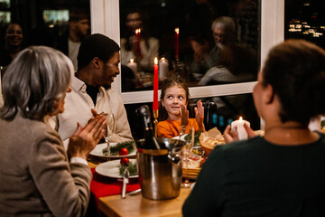 A diverse family gathers around a candlelit dinner table. A little White girl claps excitedly. An adult Black man smiles and looks at the girl and an elderly White woman with gray hair claps.