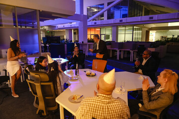A diverse group of adult colleagues gathered in an office hall after work, having a celebratory party and chatting, as a middle-aged White businessman stands next to them