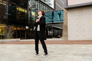 Fototapeta premium In the afternoon in a square with office buildings, a smiling and handsome dark-haired European man in his early 30s in glasses with white headphones in a black coat stands and looks up.