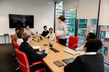 A serious dark-haired European man in his early 20s in glasses in a gray sweater discussing documents with a group of multi-ethnic colleagues sitting at a table in a meeting room during the daytime.