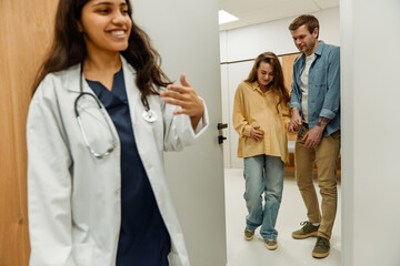 Fototapeta premium A young Indian female doctor aged 25, wearing a white doctor's coat, leading a wholesome White couple in their 30s, a pregnant wife and her husband, out of her office into the hospital hallway