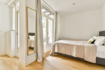 A stylish and bright bedroom featuring a large mirror, soft bedding, and natural light from large...