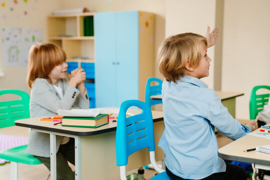 Two little White boys sit at their desks during a lesson. One boy in a blue shirt raises his hand to answer a question while looking at the teacher. The other boy looks to the right with a smile.