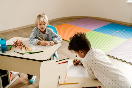 A school-aged boy with short blonde hair and a school-aged boy with brown curly hair are sitting at desks and drawing on pieces of paper during a lesson in a classroom with a multicolored carpet - Powered by Adobe