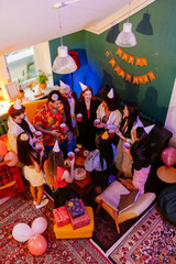 A top view of a multiethnic group of friends in their 20s partying in a living room in party hats,...