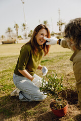 Fototapeta premium An elderly White couple plants outdoors. The smiling woman kneels by a small tree sapling while the man playfully touches her nose. They enjoy a lighthearted moment during their gardening.