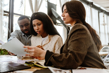 A White businesswoman in her early 30s in a black blazer sits at a table and consults a Black couple in their 20s wearing shirts and looking at documents in a building with tables and glass walls.