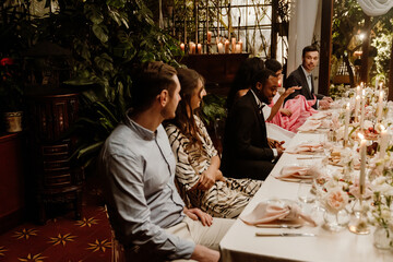 A side view of a multiethnic group of guests and a Black marrying couple in their 20s sit at a long, candlelit wedding table decorated with flowers and food in a plant-filled conservatory, talking.