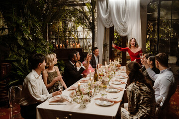 A European woman aged 30 standing up from her seat and making a toast as she's attending a wedding dinner, with guests and the young Black bride and groom sitting at the table