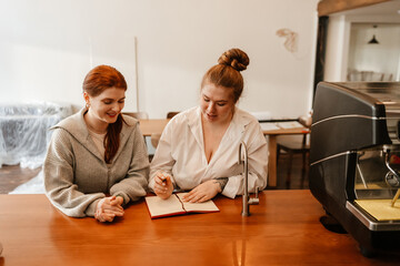 Fototapeta premium A red-haired female coffee shop owner in her early 30s wearing a white shirt talking to a happy red-haired 20-year-old friend with white skin and writing something in a notebook.