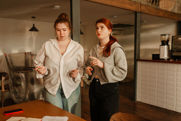 Two young White women with red hair stand discussing plans in a cafe. One gestures while speaking, and the other listens intently, holding a pen while organizing the space.