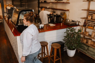 A young White female cafe owner wearing jeans and a white shirt walks past wooden stools toward the counter in a cozy coffee shop. An adult White male barista works behind the counter.