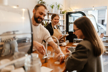 Two White baristas, a smiling man and a laughing woman, assist a young Caucasian female customer at the wooden counter of a modern coffee shop. One pours water and the other waits.