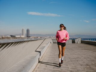 On a sunny waterfront promenade, a smiling White woman in her 20s wearing a pink sweatshirt, black shorts, white socks, sneakers, and sunglasses jogs, with a sea, city, and hills in the background.