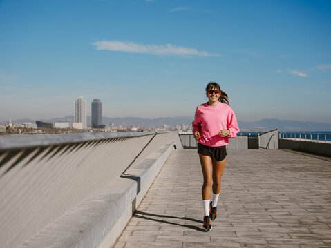On a sunny waterfront promenade, a smiling White woman in her 20s wearing a pink sweatshirt, black shorts, white socks, sneakers, and sunglasses jogs, with a sea, city, and hills in the background.