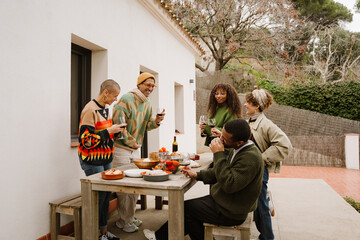 Five diverse friends socialize at an outdoor table beside a white building. Standing and sitting with glasses of red wine, they laugh and engage in conversation.