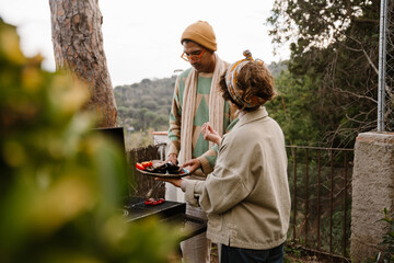 A dark-haired adult woman with white skin holds a plate of vegetables and talks to a Black man in his early 30s in a yellow hat flipping on a grill outdoors with mountains in the background.