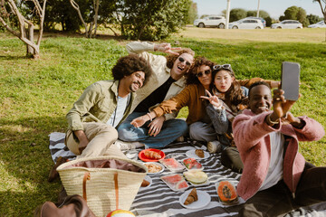 An African American man aged 25 with short, curly brunette hair, taking a selfie with a diverse group of his young friends, as they're having a picnic on a sunny day in a lush park