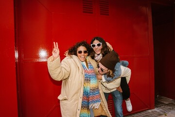 Three energetic young friends pose joyfully against a red wall. A Latino woman with curly hair shows a peace sign while a White man gives her friend a piggyback ride outdoors.