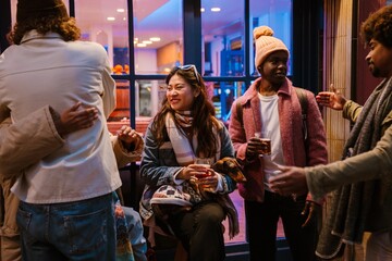 Diverse friends chat outside a cafe at night. An Asian woman with white glasses sits holding her dog and a drink, smiling while her Black, White, and Latino friends socialize nearby.