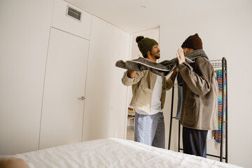 An adult gay couple standing in a bedroom next to a clothes hanger stand and dressing up, a Black man in a beanie and a jacket holding a scarf tied around a European boyfriend in a beanie and a jacket