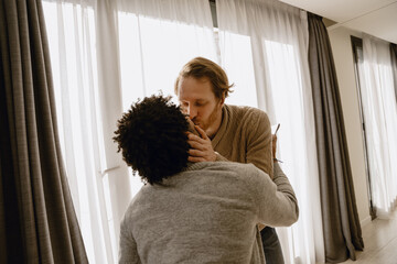 In a living room with large windows, a gay couple in their 30s, a getting up Black man with an afro wearing a grey sweater, and a White man with brown hair wearing a beige sweater, stand and kiss.