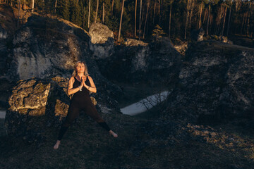 A woman practicing yoga at sunset in nature, barefoot and dressed in leg warmers and a hat,surrounded by rocks and forest. The scene reflects meditation, health, inner balance, and harmony with nature