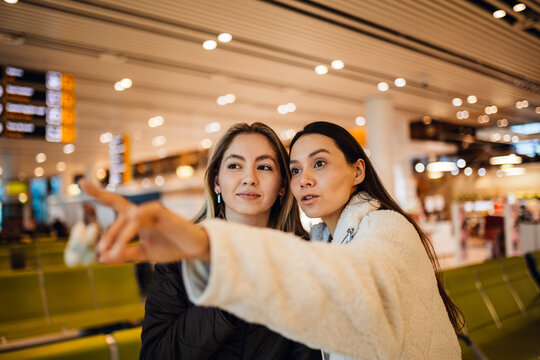Two women, a White brown-haired and an Asian brunette in their 20s, stand closely in an airport terminal, the Asian woman in a white jacket points forward while the White woman in a black jacket looks